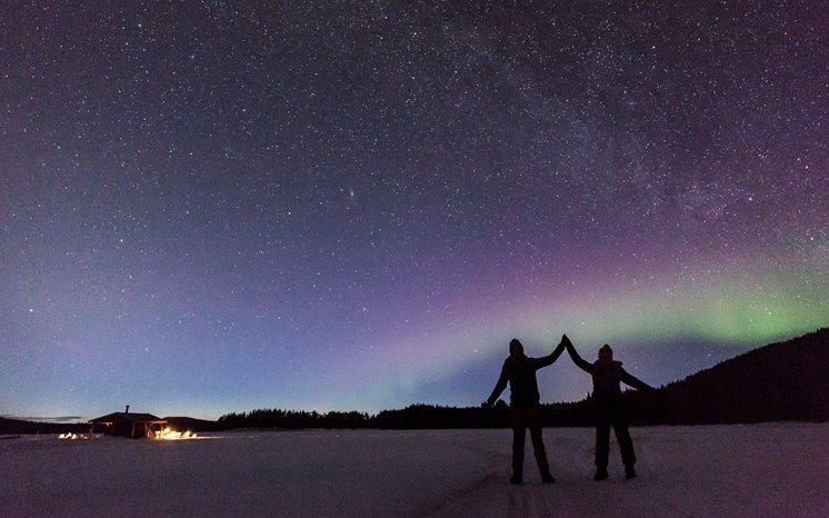 Två människor håller varandra i händerna och sträcker upp dem mot stjärnhimlen och norrskenet.
